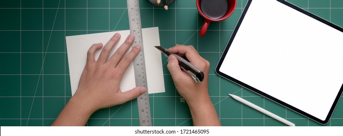 Overhead Shot Of Student Cutting Paper  With Cutter On Cutting Mat With Mock-up Tablet And Coffee Cup