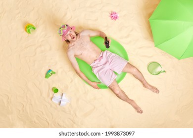 Overhead Shot Of Stunned Young Man Poses On Swim Ring At Seaside Surrounded By Beach Accessories Drinks Beer Wears Sunhat Snorkeling Mask And Shorts Tans In Sunlight. Summer Vacation Concept