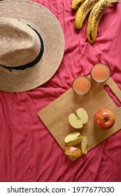 Overhead Shot Of Summer Breakfast With Apples, Bananas, Grapefruit Juice And Straw Hat, Coral Red Fabric Background, With Copy Space