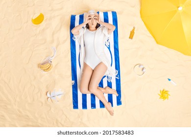 Overhead Shot Of Surprised Young Asian Woman Makes Binoculars With Hands Over Eyes Wears Swimhat And White Bikini Gets Suntan At Beach Poses On Blue Striped Towel Surrounded By Different Items