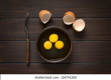 Overhead shot of three raw eggs in rustic bowl with fork on the side and broken eggshells above, photographed on dark wood with natural light - Powered by Shutterstock