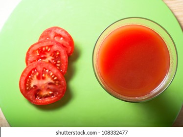 Overhead Shot Of Tomato Juice In A Long, Tall Glass Beside Three Slices On A Green Circular Mat, Resting On   Wood Table.