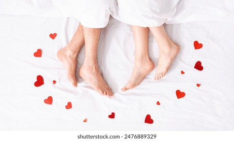 An overhead shot of two bare feet resting in a white bed with red paper hearts scattered around them. The feet are presumably from a couple in bed together, suggesting a romantic setting. - Powered by Shutterstock