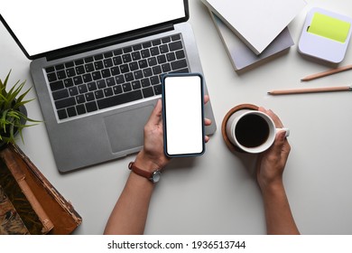 Overhead Shot View Of Woman Hand Holding Coffee Cup And Using Smart Phone At Her Office Desk With Laptop Computer.