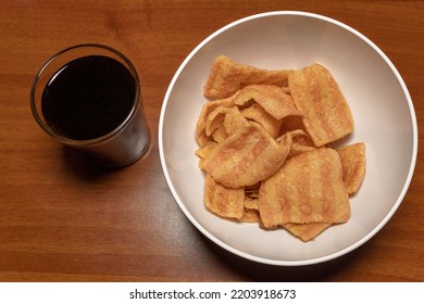 Overhead Shot Of A White Bowl Of Bacon Chips Next To A Glass Of Black Soda On A Wooden Table