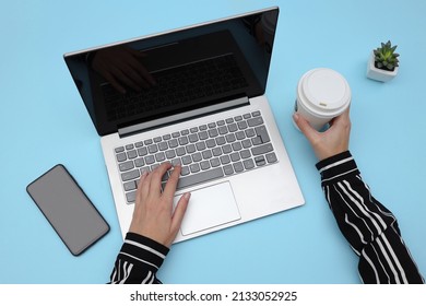 Overhead Shot Of Woman Working On Laptop While Another Hand Holding Coffee Cup, With Mobile Phone And Plant Beside On Table. Smartphone And Laptop With Blank Screen For Advertisement.