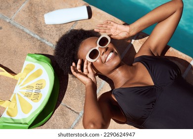 Overhead shot of young Black woman lying next to pool with purse, sunscreen - Powered by Shutterstock