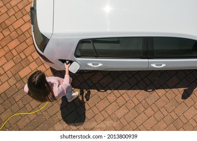 Overhead Shot Of A Young Caucasian Woman On The Electric Cars Charge Station At Daytime, Plugging In The Charger In A White Electric Car