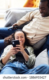 Overhead Shot Of Young Mixed Ethnicity Couple Relaxing On Sofa At Home With Man Watching Tv Whilst Woman Lies Across Lap And Checks Social Media On Mobile Phone