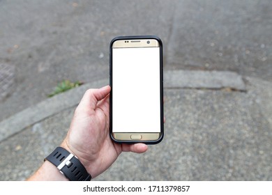 Overhead Of A Smartphone In A Business Person's Hand With A Blank Screen Who Is Stopped On A Street Corner In A Big City In Order To Read His Cellphone