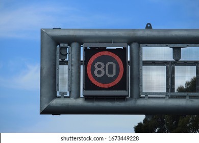 Overhead Speed Led Sign On A Bridge Over A Motorway (80 Kilometres Per Hour) Electronic 80 In A Red Circle Speed Sign Hanging Over A Freeway. Blue Sky Background.