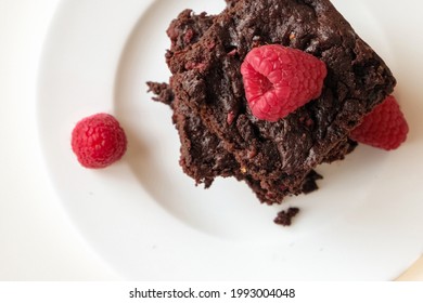 Overhead Of Stack Of Dark Chocolate Brownie With Raspberries On A White Plate Against Light Background