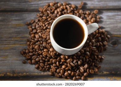 Overhead studio photograph of a white patterned cup filled with freshly brewed black coffee, surrounded by a scattered pile of roasted coffee beans on a rustic wooden table texture.