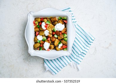 Overhead Of Tater Tot Southwestern Casserole Topped With Fresh Avocado, Tomatoes, And Dollops Of Rich Sour Cream In Square, Southern Casserole Dish On Turquoise Striped Linen On Cream Marble Surface.