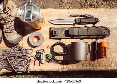 Overhead top view of hiking travel gear on wood log. Items include hiking boots, cup, rope, knife, matches, flashlight, compass. Flat lay of outdoor travel equipment items for mountain camping trip.