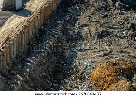 Overhead view of active construction site featuring deep excavation trench, sturdy concrete pile retaining wall, and rough terrain with distinct pile of orange sand under bright sunlight.