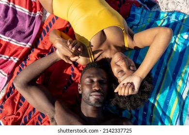 Overhead View Of African American Couple Holding Hands While Lying Together On Beach Towel. Unaltered, Love, Lifestyle, Togetherness, Enjoyment And Holiday Concept.