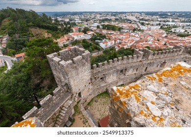 Overhead view of an ancient stone fortress with battlements, overlooking a charming town with red-tiled rooftops, lush greenery, and a dramatic cloudy sky - Powered by Shutterstock