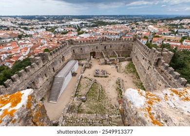 Overhead view of an ancient stone fortress with battlements, overlooking a charming town with red-tiled rooftops, lush greenery, and a dramatic cloudy sky - Powered by Shutterstock