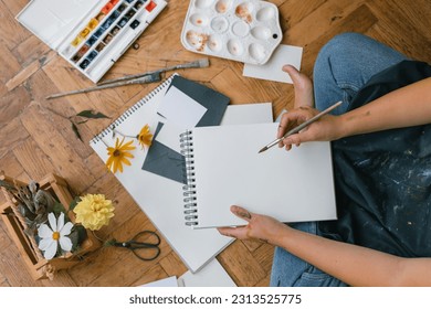 Overhead view of artist in apron sitting on the floor and painting with watercolour paints. Art supplies on the floor. Woman holding blank sketchbook and paint brush in her hands. - Powered by Shutterstock