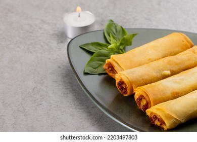 Overhead View Of Asian Spring Rolls On Grey Plate And Lit Tea Candle On Grey Background. Asian Tasty Home Cooked Food And Healthy Eating.