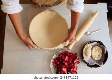 Overhead view of a baker shaping dough for a homemade pie with fresh plums on a kitchen counter. Rolling pin and ingredients are visible, emphasizing a rustic baking process. - Powered by Shutterstock