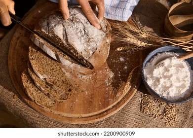 Overhead View Of A Baker Slicing Whole Grain Bread On A Cutting Board With A Kitchen Knife. A Bowl Of Flour And Spikelets Of Wheat, Crumbs And Wheat Grains Are Scattered On The Burlap Tablecloth