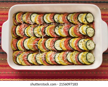 Overhead View Of A Baking Dish Of Alternating Zucchini, Yellow Squash, And Tomato Circles, Topped With Parmesan Cheese And Garnished With Fresh Rosemary And Thyme, Against A Striped Table Covering