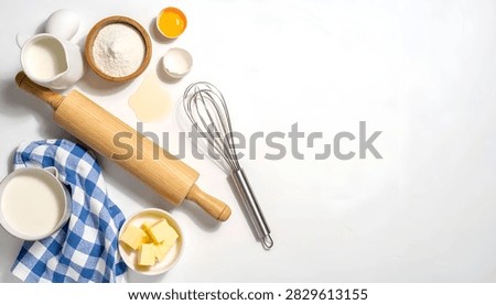 Overhead view of baking essentials including flour, butter, eggs, milk, a rolling pin, and a whisk, laid out on a clean white background.