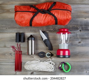 Overhead View Of Basic Hiking Gear Placed On Weathered Wooden Boards. Items Include Tent Inside Of Bag, Pegs, Compass, Canteen, Rope, Knife, Case, Lantern And Binoculars. 