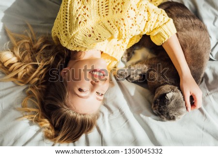 overhead view of beautiful smiling girl stroking scottish fold cat while lying in bed at home