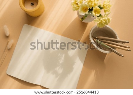 Overhead view of a blank note book, stationery and flowers in vase on a table in sunlight.