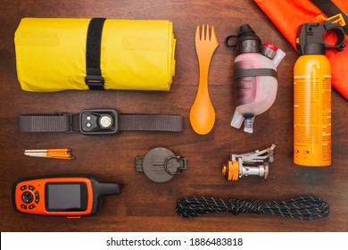 Overhead view of camping gear and equipment including headlamp, GPS, compass, rope, bear spray, water filter, and sleeping pad on a wooden background