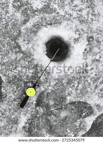 An overhead view captures a circular hole in thick, textured ice of a frozen lake, where a bright yellow fishing reel and rod extend into the dark water. This winter fishing scene highlights the contr