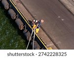 An overhead view captures two dock workers in hard hats and safety vests actively engaged in mooring operations at a port. They are shown securing thick ropes around bright yellow bollards on the concrete dock. Along the dock wall, several large tire fenders hang, providing protection. The dark green water of the harbor is visible next to the dock, which features a yellow and black striped safety edge. In the background, a paved area with industrial tracks suggests further port infrastructure, and a red and white striped warning marker is on the ground. The scene depicts typical maritime activity in Gdynia, Poland.