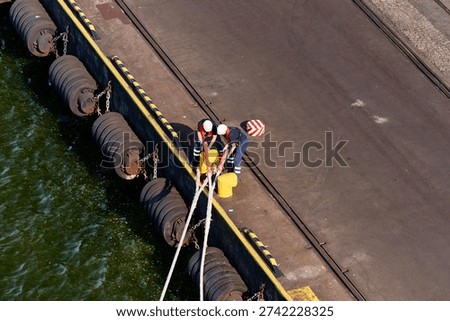 An overhead view captures two dock workers in hard hats and safety vests actively engaged in mooring operations at a port. They are shown securing thick ropes around bright yellow bollards on the concrete dock. Along the dock wall, several large tire fenders hang, providing protection. The dark green water of the harbor is visible next to the dock, which features a yellow and black striped safety edge. In the background, a paved area with industrial tracks suggests further port infrastructure, and a red and white striped warning marker is on the ground. The scene depicts typical maritime activity in Gdynia, Poland.