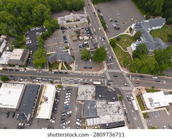 Overhead view capturing a bustling suburban town center with an active intersection, numerous vehicles driving and parked, commercial buildings, a church, and surrounding green trees
