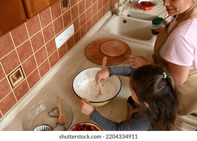 Overhead View: Charming Little Girl Standing By Countertop, Kneading Dough For Delicious Festive Cherry Pie, Helping Her Mother In The Kitchen. Child Learns Culinary. Mom And Daughter Cooking Together