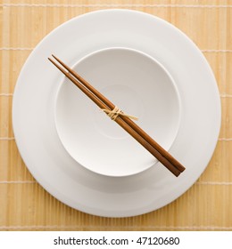 Overhead View Of Chopsticks Lying Across An Empty Bowl That Is Sitting On A Plate On A Bamboo Mat. The Dishes Are White. Square Shot.