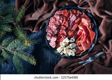 Overhead View Of A Christmas Antipasti - Cheese, Sausages And Dry Cured Meat - Jamon,  Chorizo, Blue Mold Cheese, Sliced Fuet Sausages On A Plate On A Table With Fir Tree, View From Above, Flatlay