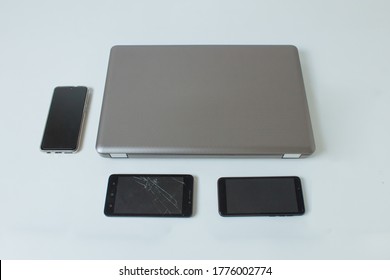 An Overhead View Of A Closed Gray Laptop On A White Table Surrounded By Three Mobile Phones, One Of Them With A Broken Screen Next To It.