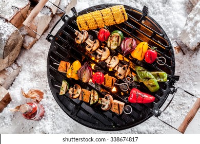 Overhead view of colorful vegetable kebabs and a corncob grilling on a winter BBQ outdoors in snow with tasty spicy dips and the wood pile alongside