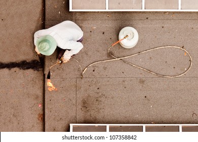 Overhead View Of Construction Worker Using Propane Torch With Gas Cylinder To Patch Tar Paper On Flat Roof