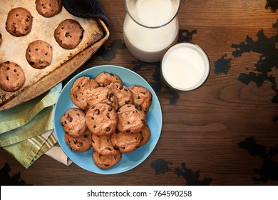 Overhead View Of Cookie Sheet And Plate Of Chocolate Chip Cookies Warm From The Oven With Glass Of Cold Milk On Vintage Wood Table