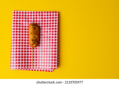 Overhead View Of Corn Dog With Mustard Sauce On Checked Pattern Napkin Over Yellow Background. Unaltered, Copy Space, Unhealthy Food, Meat, Sausage And Fried Food.