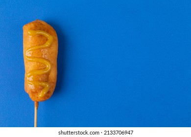 Overhead View Of Corn Dog With Mustered Sauce Over Skewer On Blue Background With Copy Space. Unaltered, Copy Space, Unhealthy Food, Meat, Sausage And Fried Food.