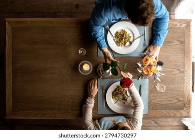 Overhead view of a couple dining at a rustic wooden table, sharing pasta and flowers in a cozy romantic dinner setting.