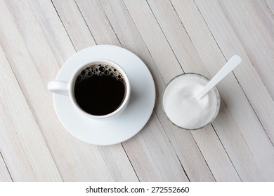 Overhead View Of A Cup Of Coffee And A Bowl Of Granulated Sugar With A Plastic White Spoon. Horizontal Format On A Rustic Whitewashed Table.