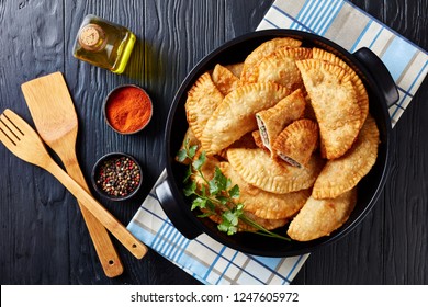 overhead view of deep fried turnovers or chebureki with a filling of ground beef meat and onion in a black ceramic pan on a wooden table, view from above, close-up, flatlay