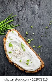 Overhead View Of Delicious Cream Cheese And Chopped Chives On A Slice Of Rye Or Wholewheat Bread On A Dark Textured Surface With Copyspace